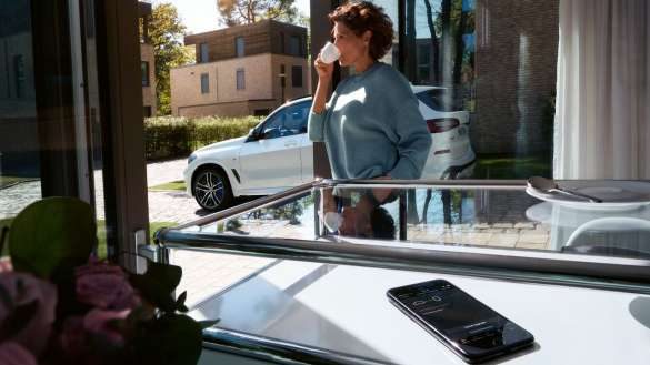 BMW 3er Limousine M Automobile G80 G20 Remote Software Upgrade Fokus auf Smartphone im Vordergrund mit Frau und BMW im Hintergrund